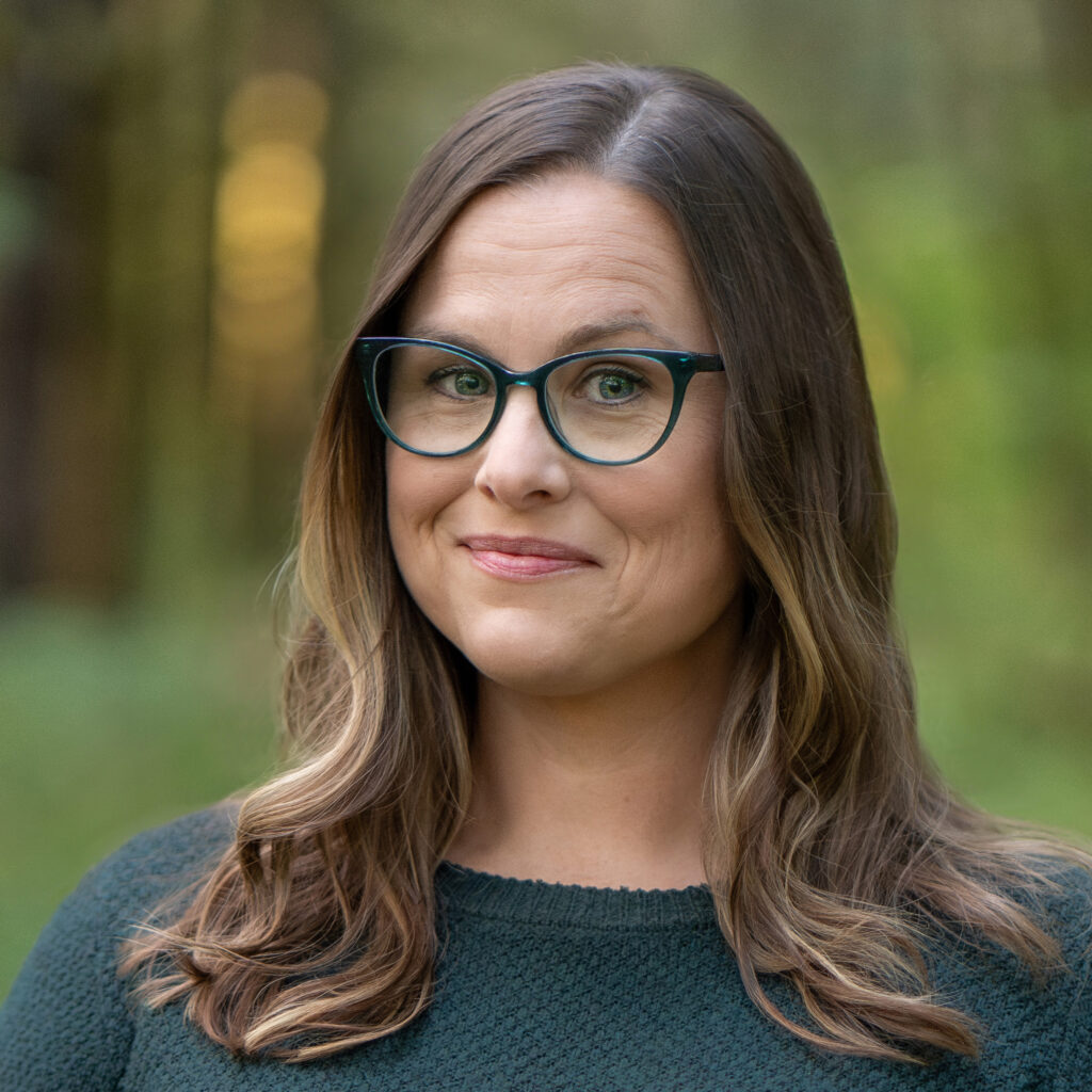 Sarah is a woman with light skin, long brown hair with highlights, and green-framed glasses. She is standing in a forest wearing a dark green sweater and smiling slightly.